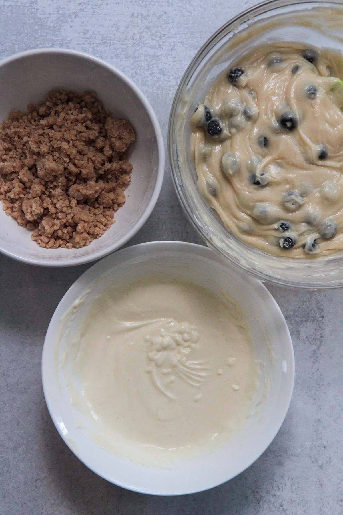 three separate bowls filled with muffin batter, cream cheese filling, and streusel topping. 
