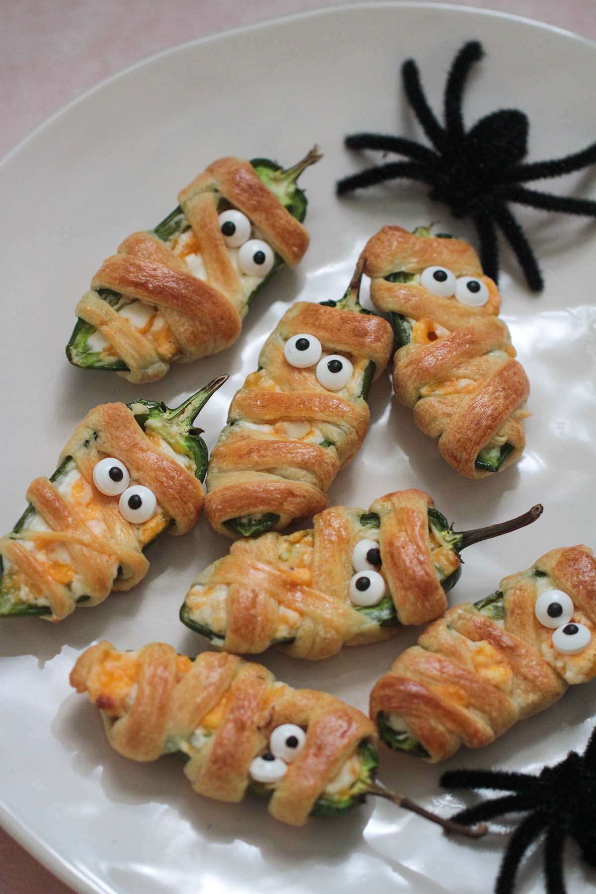 seven mummy jalapeno poppers on a white plate with 2 fake spiders.