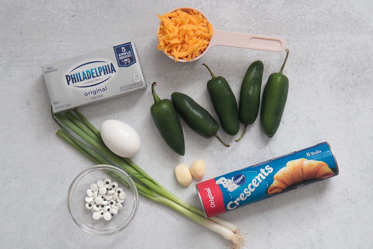 ingredients for mummy jalapeno poppers. cheese, dough, jalapenos, egg, garlic, onions, and candy eyes.