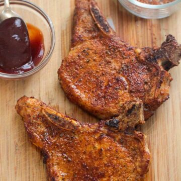two air fried bone-in pork chops on a wooden board with barbecue sauce and a spice rub on the side.
