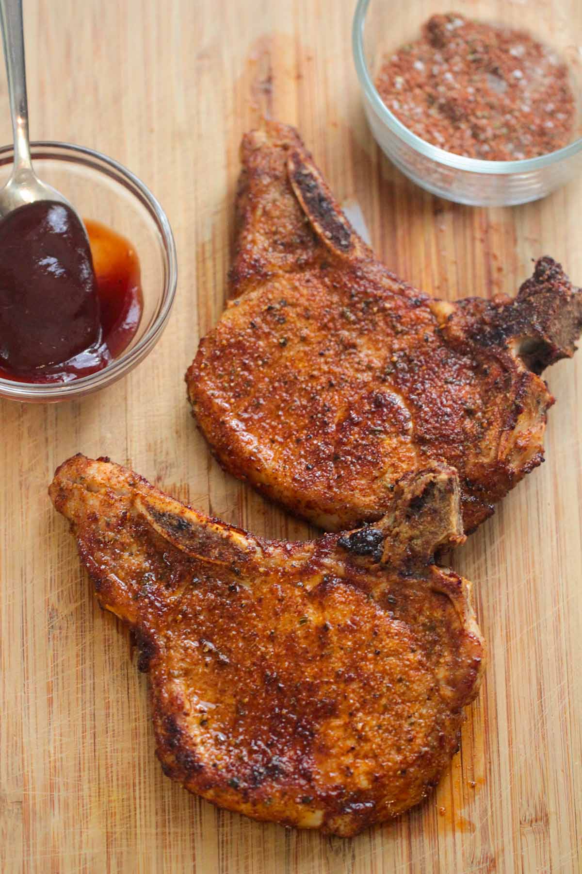 two air fried bone-in pork chops on a wooden board with barbecue sauce and a spice rub on the side. 