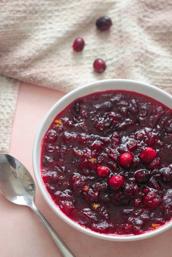 classic cranberry sauce in a bowl with a spoon, towel, and cranberries on the side.