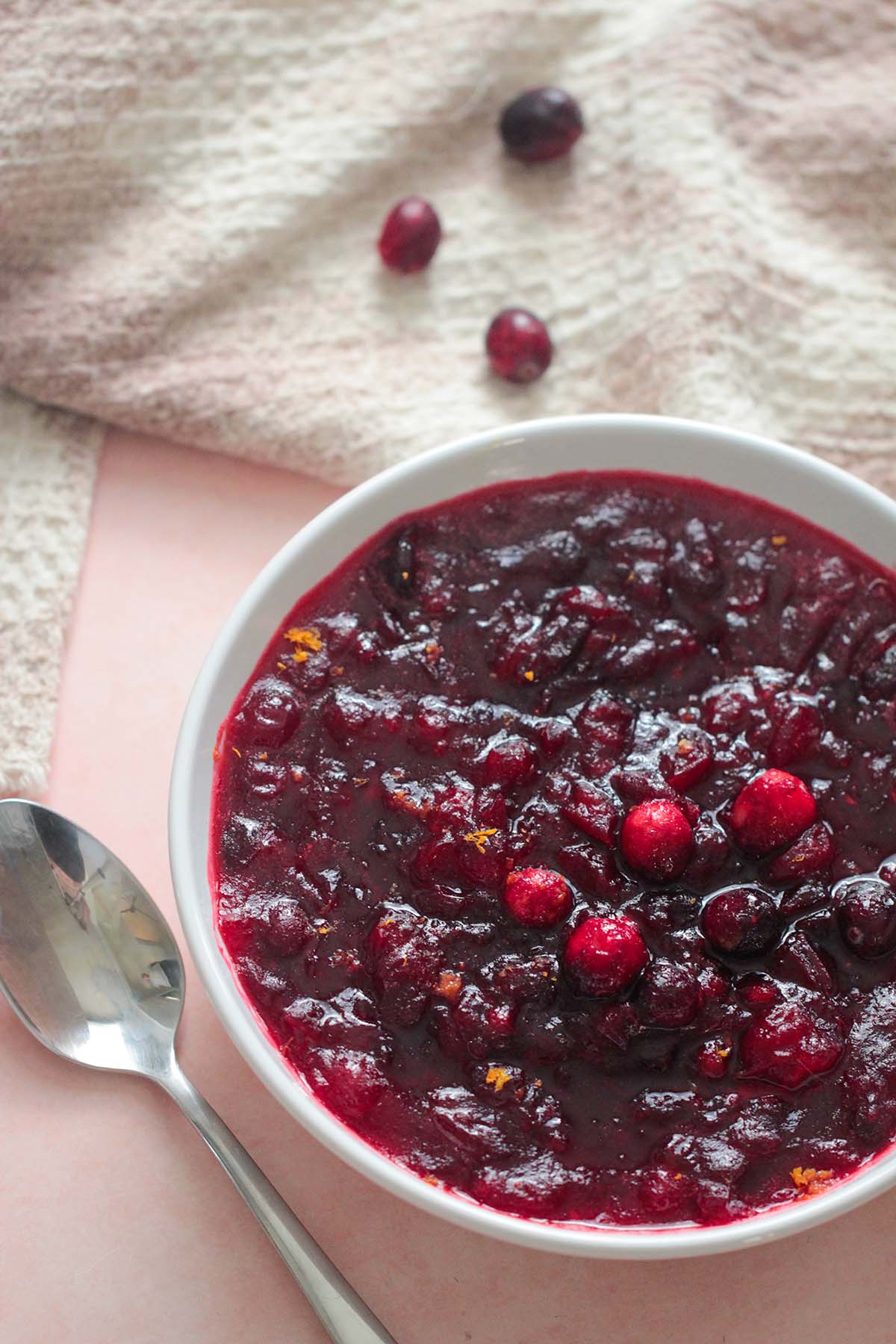 classic cranberry sauce in a white bowl with a spoon, cranberries, and towel on the side. 