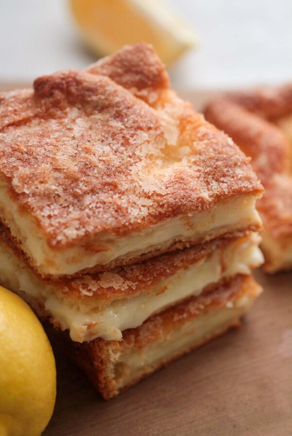 three lemon cream cheese bars stacked on a wooden board with a lemon on the side.