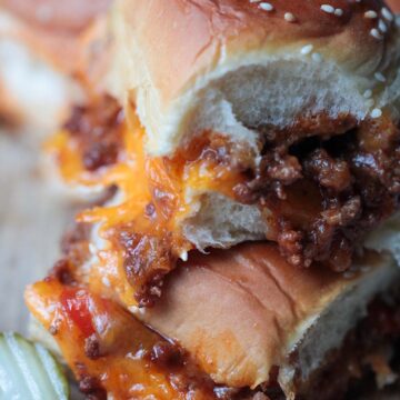 two sloppy joe sliders stacked on a wooden board with a pickle on the side.