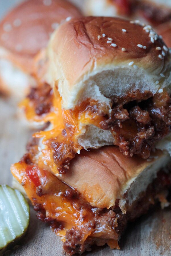 two sloppy joe sliders stacked on a wooden board with a pickle on the side.