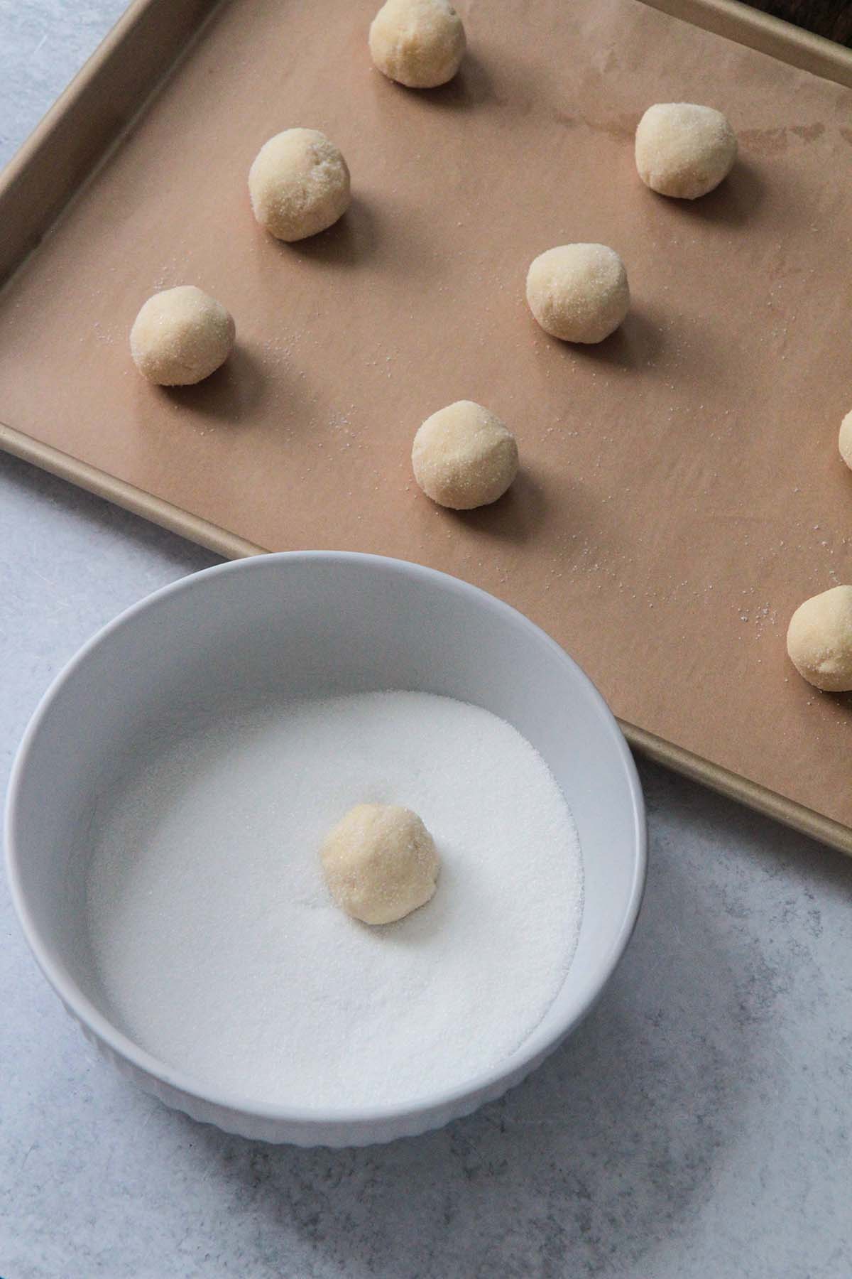 cookie dough balls on a baking sheet and a cookie dough ball in a bowl of sugar. 