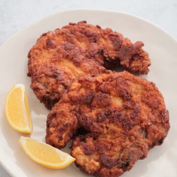two fried pork chops on a plate with two lemon wedges.