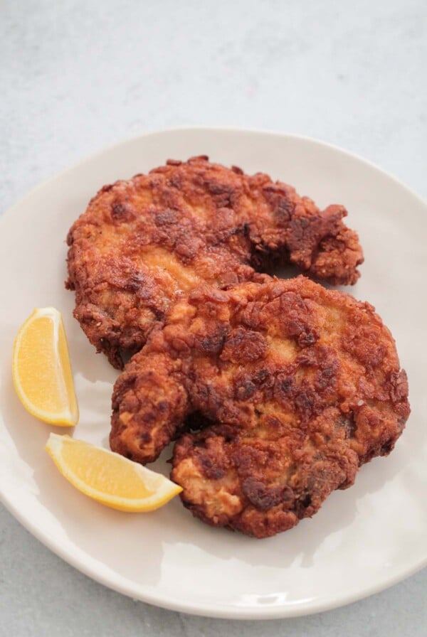 two fried pork chops on a plate with two lemon wedges.