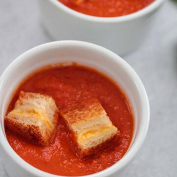 Two small bowls of creamy tomato soup with grilled cheese croutons on top.
