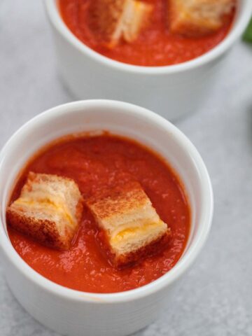 Two small bowls of creamy tomato soup with grilled cheese croutons on top.