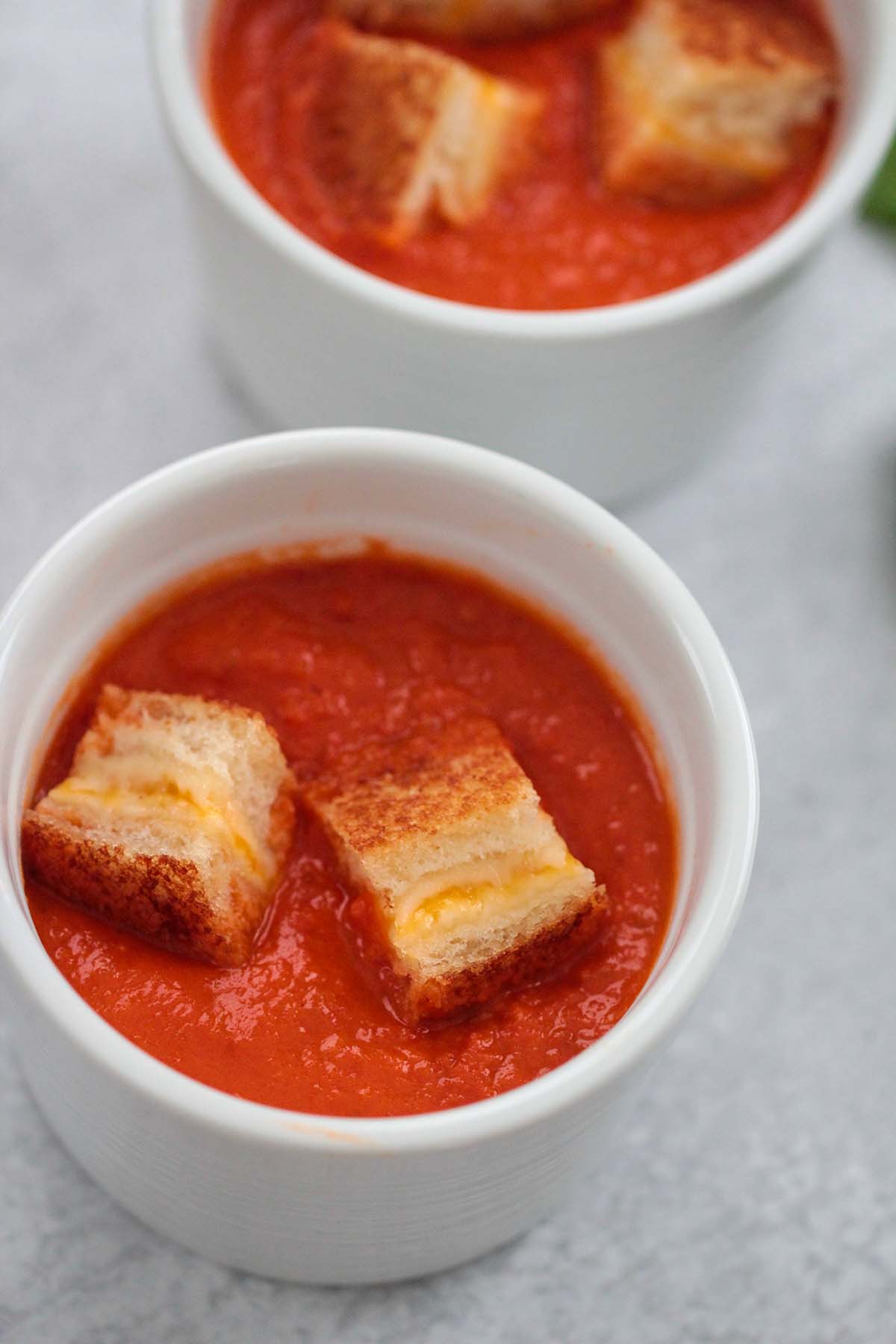 Two small bowls of creamy tomato soup with grilled cheese croutons on top. 