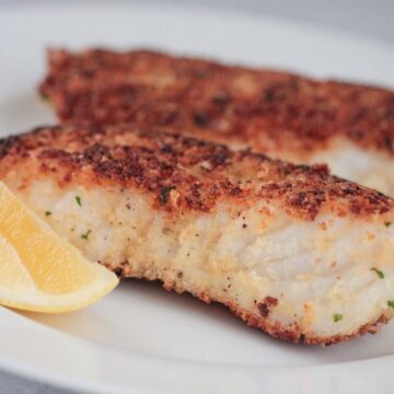 two pan-seared parmesan crusted halibut fish on a plate with a lemon wedge on the side.
