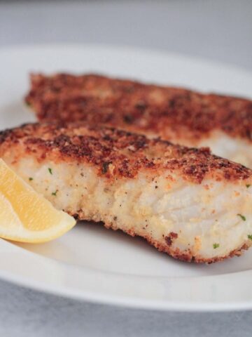 two pan-seared parmesan crusted halibut fish on a plate with a lemon wedge on the side.
