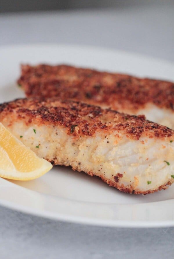 two pan-seared parmesan crusted halibut fish on a plate with a lemon wedge on the side.