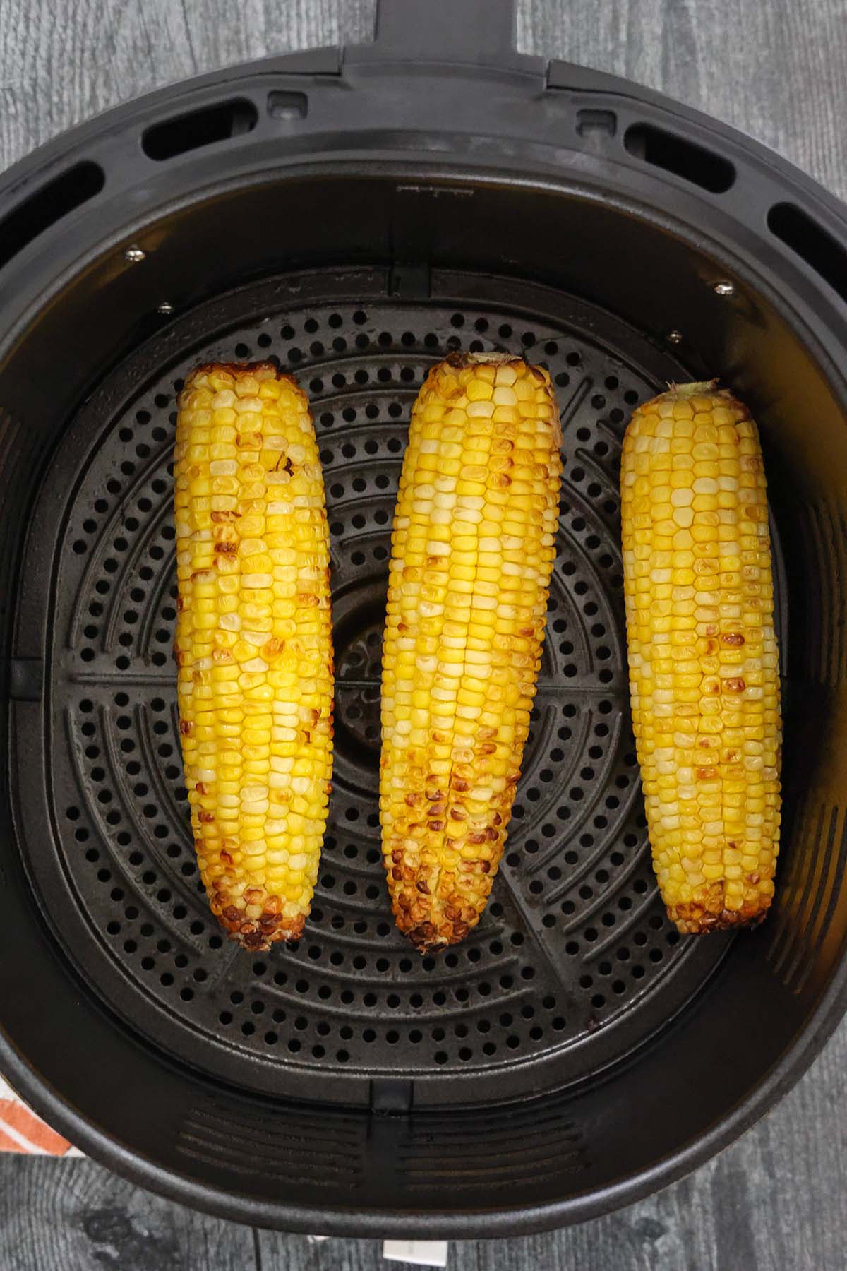 three cooked corn on the cobb in the air fryer basket. 