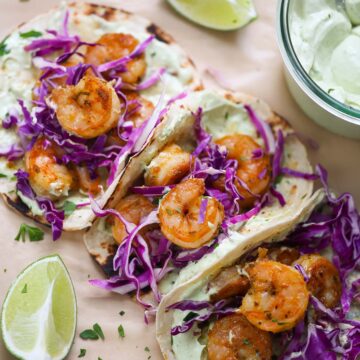 Three air fryer shrimp tacos with limes and avocado crema on the side.