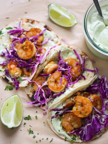 Three air fryer shrimp tacos with limes and avocado crema on the side.