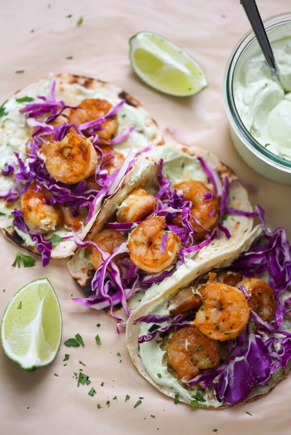 Three air fryer shrimp tacos with limes and avocado crema on the side.