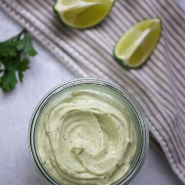 avocado crema in a jar with limes, cilantro, and avocado in the background.