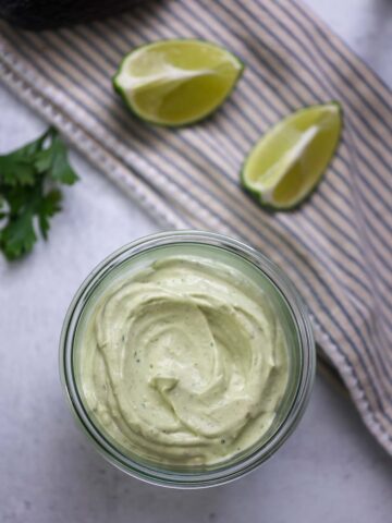 avocado crema in a jar with limes, cilantro, and avocado in the background.