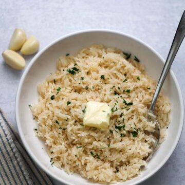 Garlic buttery rice in a white bowl with butter and parsley on top and a spoon on the side.