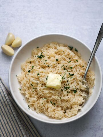 Garlic buttery rice in a white bowl with butter and parsley on top and a spoon on the side.
