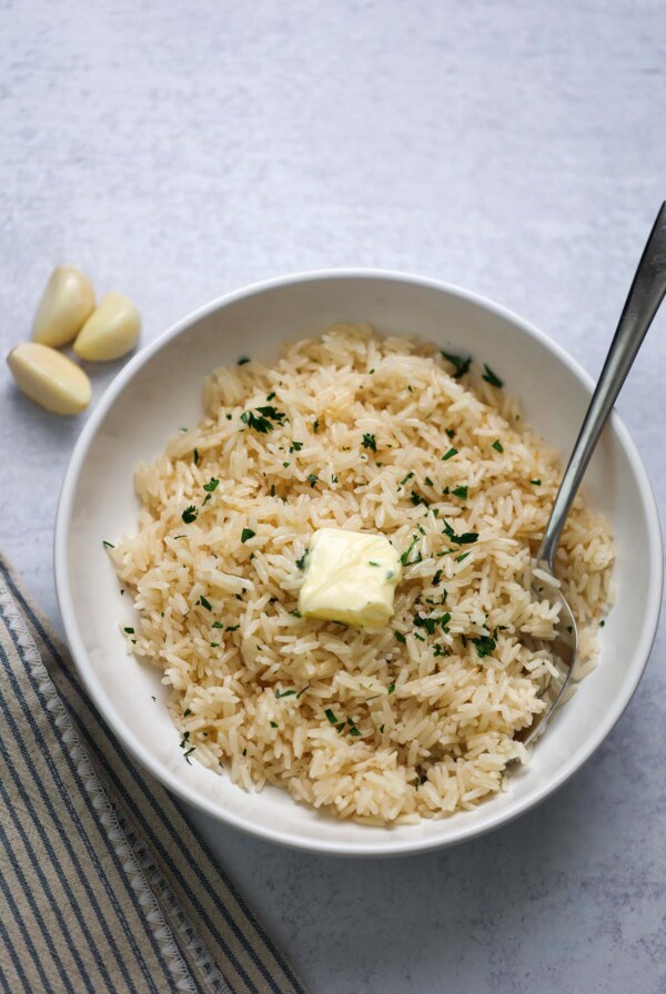 Garlic buttery rice in a white bowl with butter and parsley on top and a spoon on the side.
