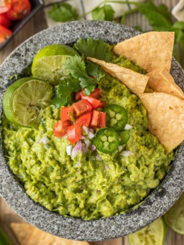 Guacamole in a molcajete topped with limes, tomatoes, jalapenos, and chips.