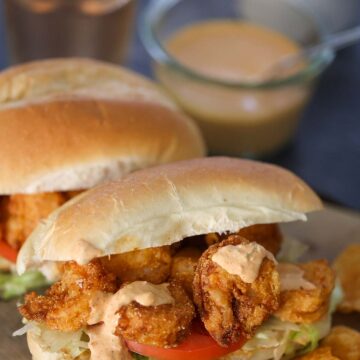 Two crispy shrimp po'boy sandwiches on a wooden board with chips and a pickle on the side.