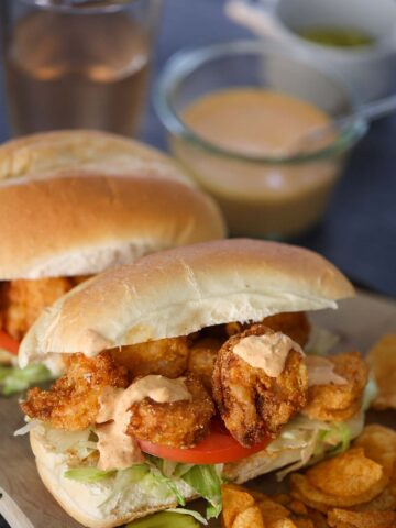 Two crispy shrimp po'boy sandwiches on a wooden board with chips and a pickle on the side.
