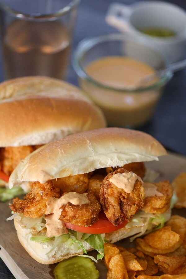 Two crispy shrimp po'boy sandwiches on a wooden board with chips and a pickle on the side.
