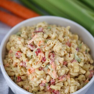 Deli-style macaroni salad in a white bowl with carrots and celery in the background.