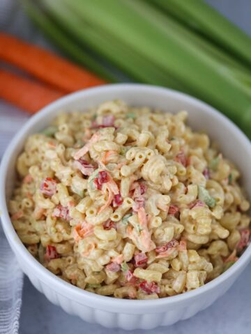 Deli-style macaroni salad in a white bowl with carrots and celery in the background.