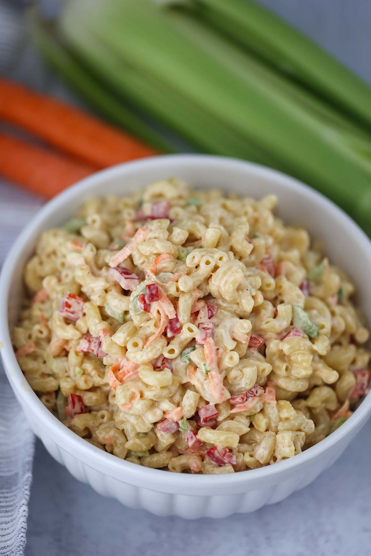 Deli-style macaroni salad in a white bowl with carrots and celery in the background. 