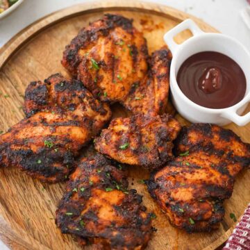 five grilled boneless chicken thighs on a wooden board with bbq sauce on the side.