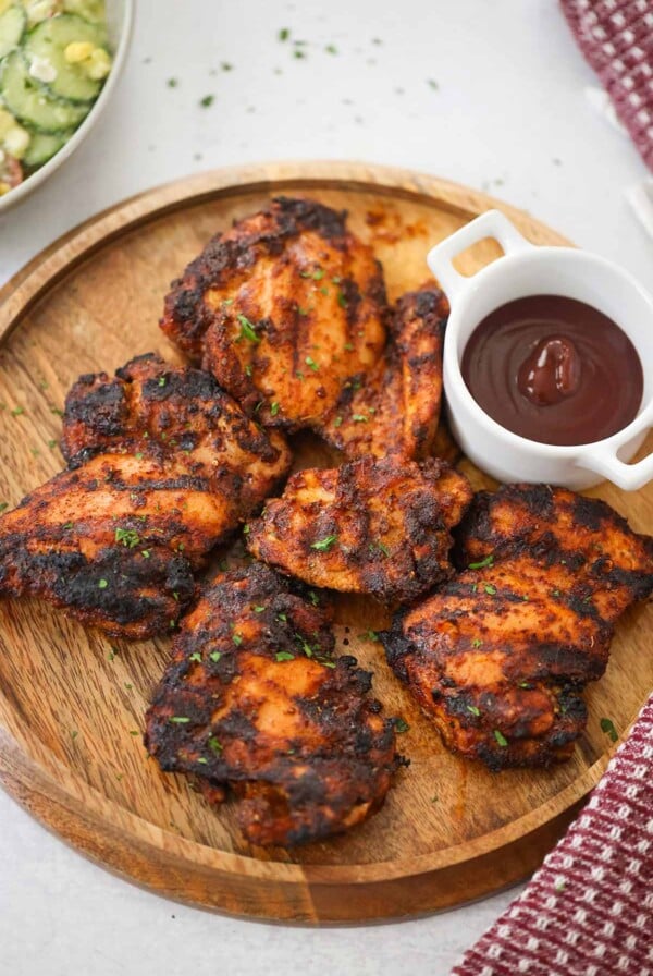 five grilled boneless chicken thighs on a wooden board with bbq sauce on the side.