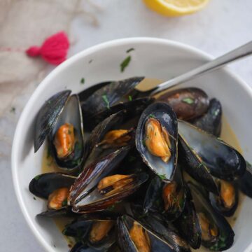 black opened mussels with a spoon in a white bowl with a lemon on the side.