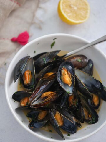 black opened mussels with a spoon in a white bowl with a lemon on the side.
