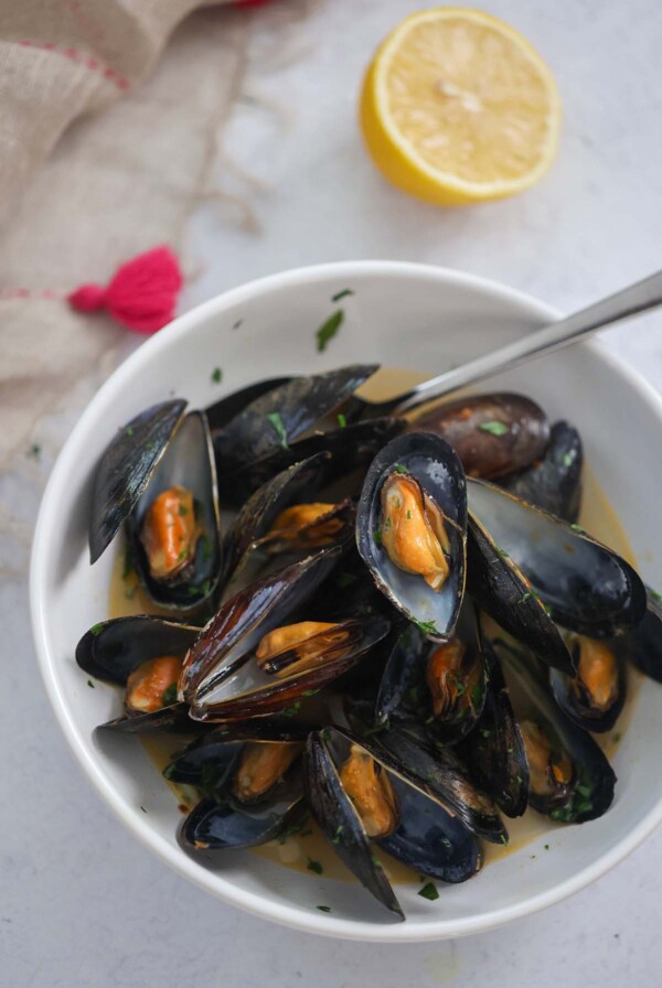black opened mussels with a spoon in a white bowl with a lemon on the side.