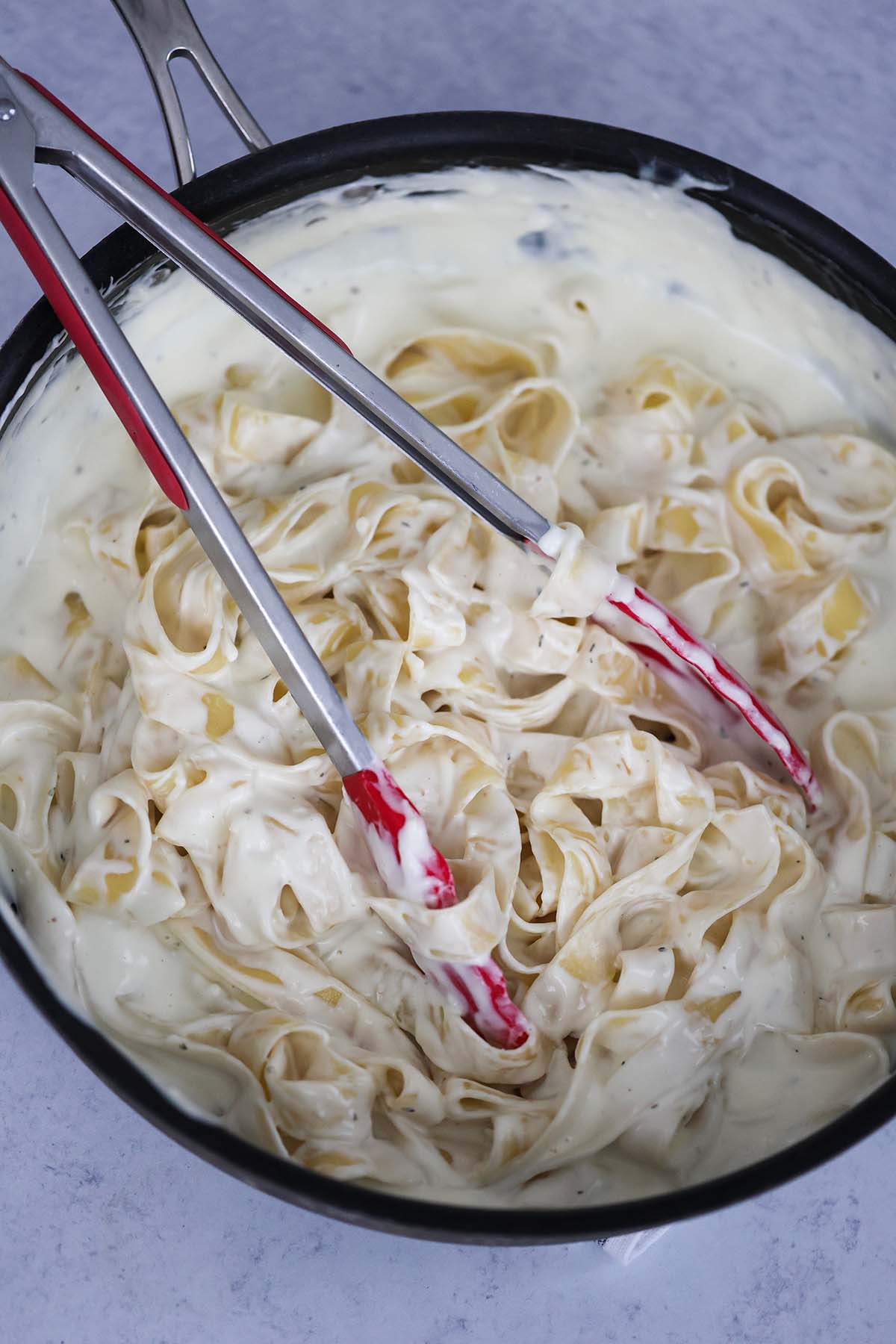 Tagliatelle coated in Alfredo sauce in a skillet with tongs. 