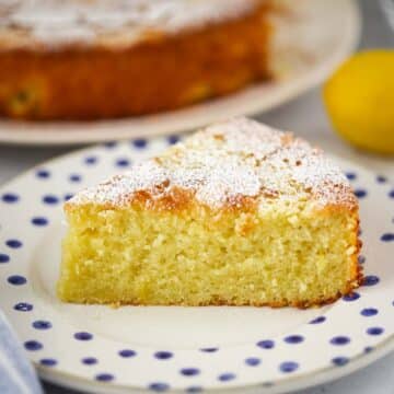 lemon olive oil cake slice on a plate with the entire cake in the background.