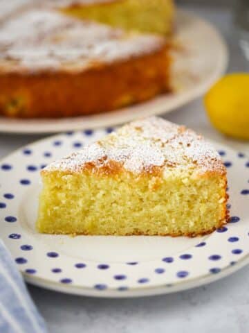 lemon olive oil cake slice on a plate with the entire cake in the background.