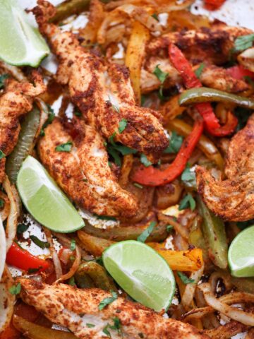 baked chicken tenders, onions, peppers, and lime wedges on a sheet pan.