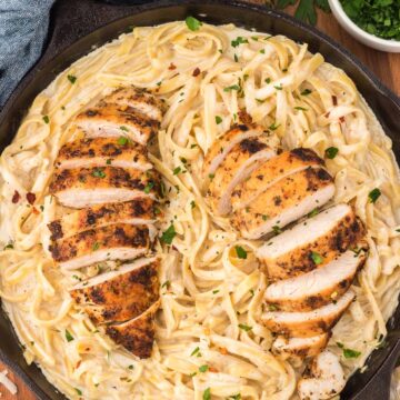 chicken fettuccine alfredo in a black skillet with red pepper flakes and parmesan cheese on the side.
