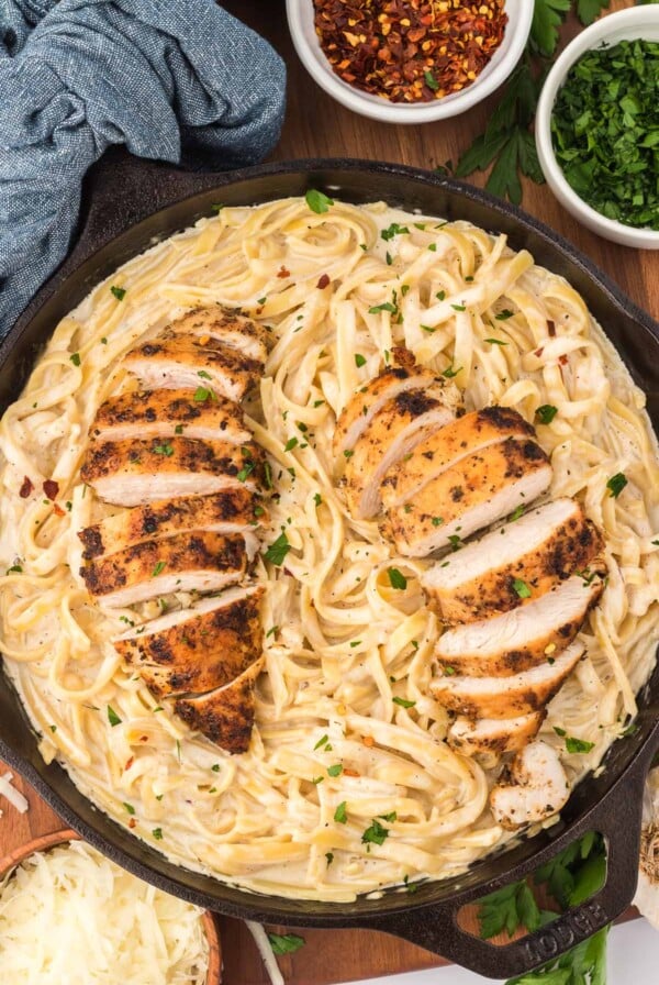 chicken fettuccine alfredo in a black skillet with red pepper flakes and parmesan cheese on the side.