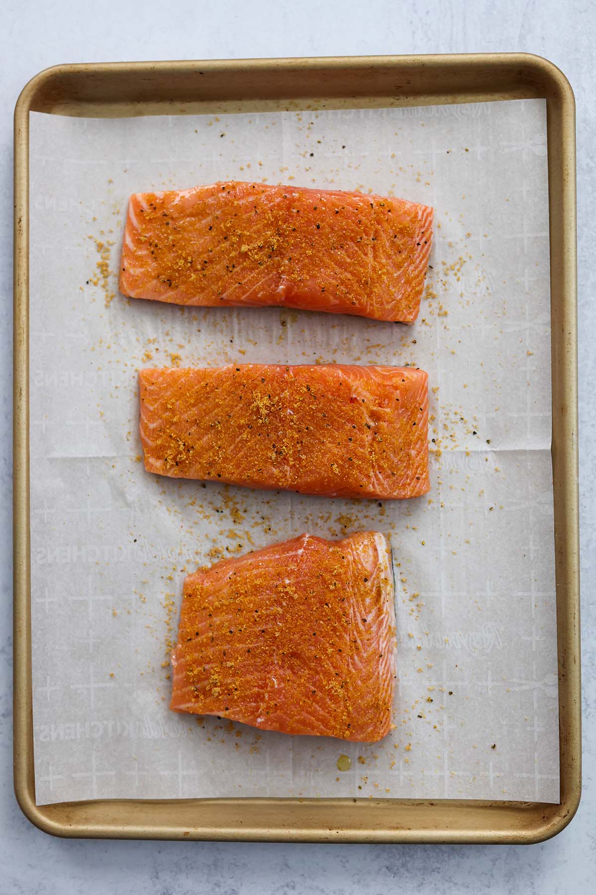 three raw salmon fillets seasoned with lemon pepper on a baking sheet. 