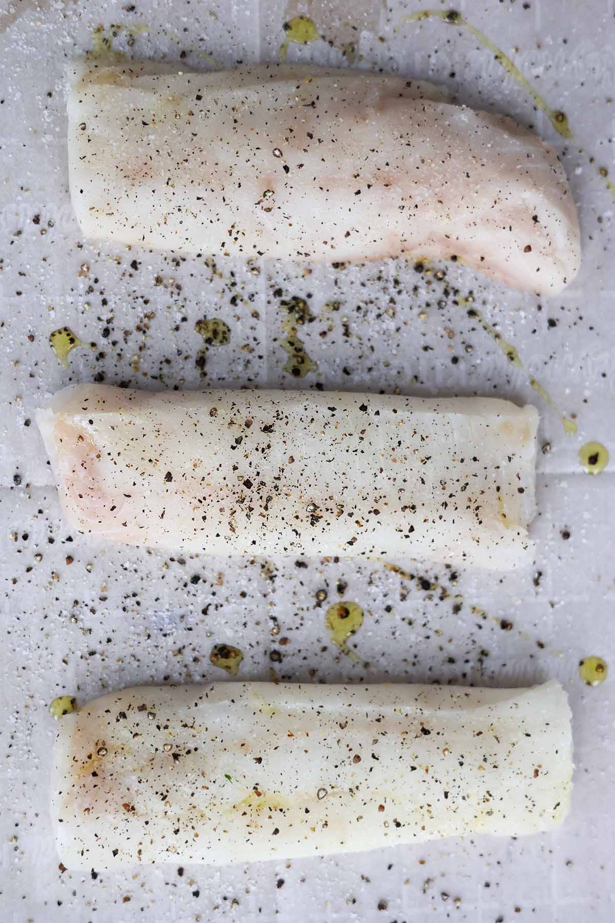 three raw cod fish fillets seasoned with salt and pepper on a baking sheet. 