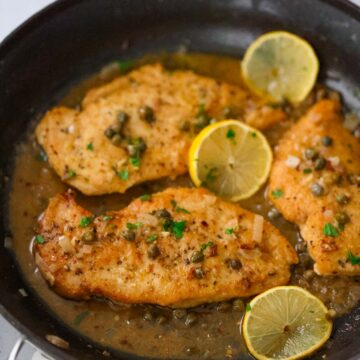 three chicken breasts piccata in a skillet with sauce and lemon slices.