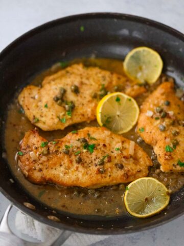 three chicken breasts piccata in a skillet with sauce and lemon slices.